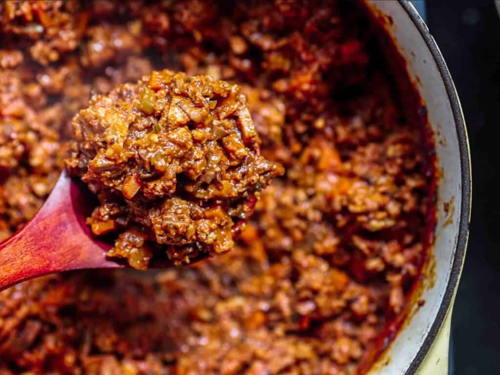 A close-up of a wooden spoon scooping hearty, chunky vegan bolognese from a pot, with visible pieces of plant-based meat and onions in a rich, red tomato base.